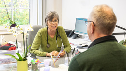 Das Bild zeigt eine freundliche Szene in einer Arztpraxis: Eine Ärztin im Gespräch mit einem älteren Patienten. Beide sitzen sich an einem Schreibtisch gegenüber. Die Ärztin trägt ein Stethoskop um den Hals und hört aufmerksam zu. Im Hintergrund ist ein Computerbildschirm zu sehen, auf dem medizinische Daten angezeigt werden.