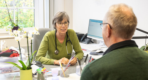 Das Bild zeigt eine freundliche Szene in einer Arztpraxis: Eine Ärztin im Gespräch mit einem älteren Patienten. Beide sitzen sich an einem Schreibtisch gegenüber. Die Ärztin trägt ein Stethoskop um den Hals und hört aufmerksam zu. Im Hintergrund ist ein Computerbildschirm zu sehen, auf dem medizinische Daten angezeigt werden.