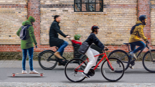 Drei Personen auf Fahrrädern sowie ein Mann auf einem Skateboard fahren auf einer Straße an der Kamera vorbei.