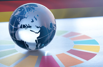 Eine Weltkugel aus Glas steht auf einem Tisch. Da drum sieht man den SDG Colour Wheel. Im Hintergrund ist die Deutschlandflagge zu sehen.