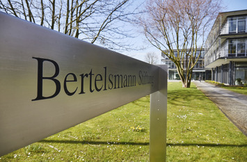 Außenaufnahmen der Bertelsmann Stiftung im Frühling. Schild mit dem Schriftzug Bertelsmann Stiftung im Vordergrund, im Hintergrund das Gebäude der Stiftung.