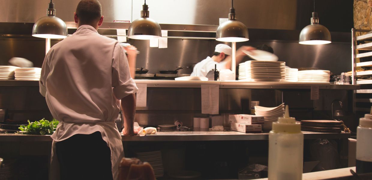 (© Michael Browning / unsplash.com (Public Domain)) Ein Koch-Azubi steht an einer Arbeitsplatte in einer Restaurant-Küche und bereitet ein Gericht zu. Er ist nur von hinten zu sehen. Im Hintergrund stehen vier Kollegen von ihm und bereiten ihre Gerichte zu, einer der Kollegen greift nach einem Teller.