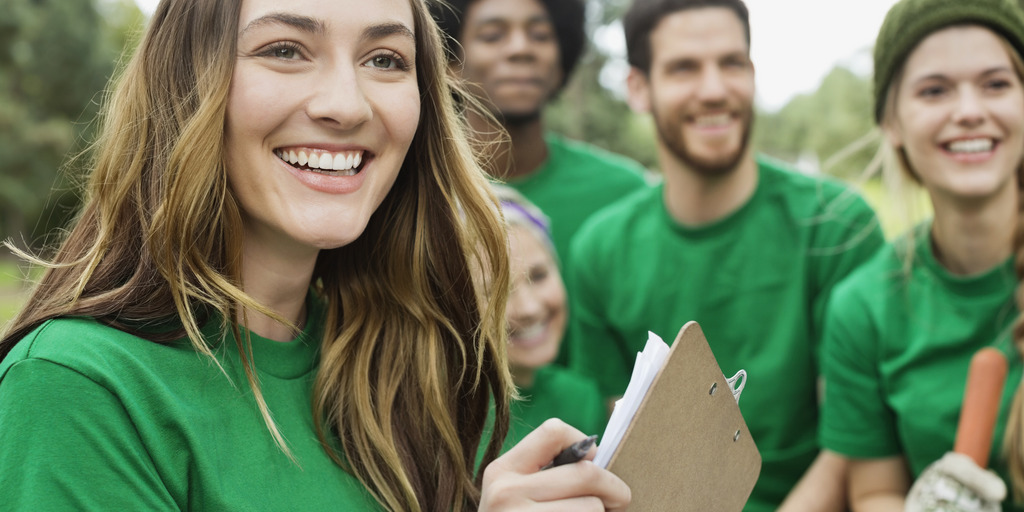 Im Vordergrund ein junges, lachendes Mädchen, im Hintergrund weitere, diverse Personen. Alle gleich gekleidet, in grün, und alle lachen.