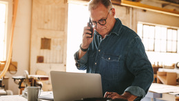 Älterer Mann steht in einer Tischlerwerkstatt an einem Arbeitstisch. Auf dem Tisch steht ein Laptop, in das er schaut, während er mit der rechten Hand telefoniert.