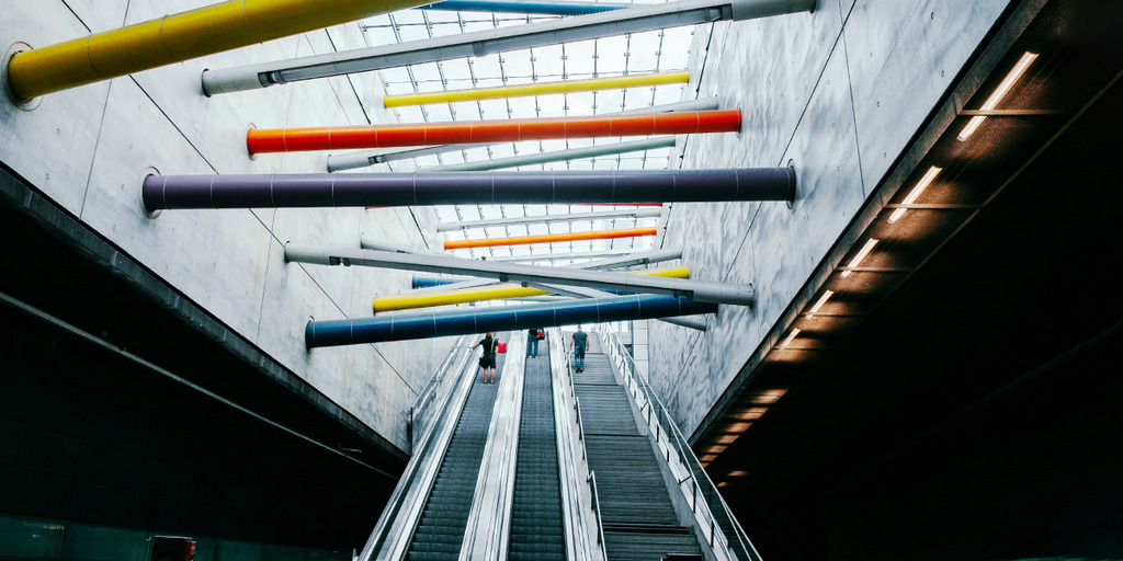 Rolltreppen in einer U-Bahn-Station. Der Blick richtet sich an die Decke wo in bunten Farben Verbindunsbalken zwischen der rechten und linken Wand zu sehen sind.