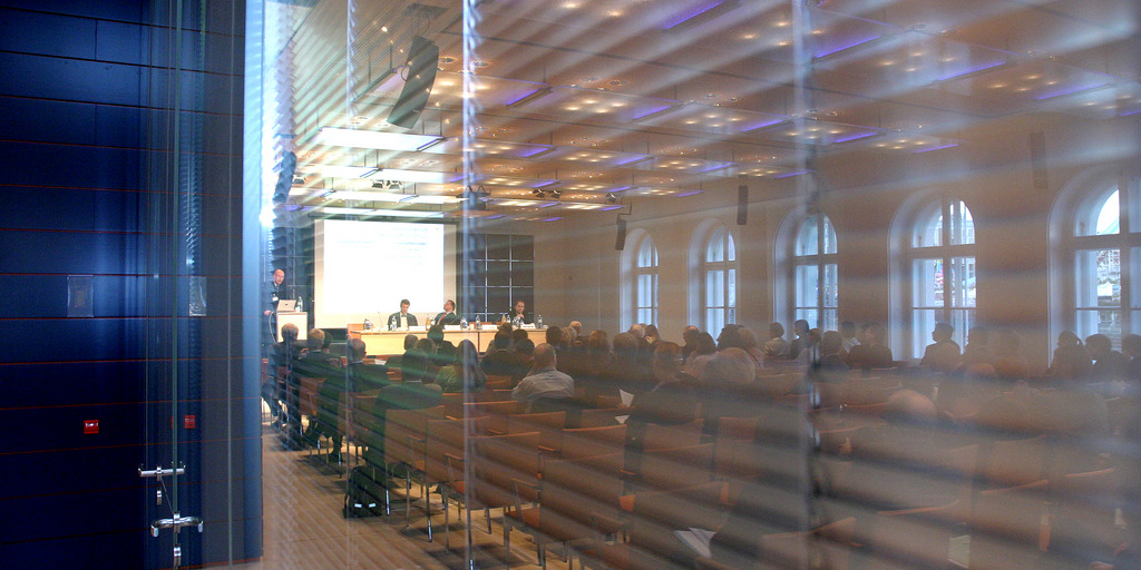 Der Blick zeigt einen Veranstaltungsraum durch eine Glaswand. Im Raum stehen Stuhlreihen.