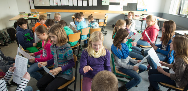 viele Kinder sitzen sich auf ihren Stühlen im Klassenraum gegenüber, sie halten ein Blatt Papier in der Hand