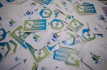 Ein ungeordneter Haufen quadratischer Aufkleber und K&auml;rtchen liegt auf einer hellen Fl&auml;che verteilt. Darauf sind gr&uuml;ne und blaue Logos sowie die Aufschriften &bdquo;Daten Dialog&ldquo;
