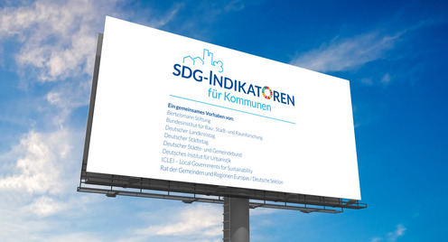 Auf dem Foto ist ein Schild mit dem Logo der SDG-Indikatoren für Kommunen zu sehen. Das Logo zeigt die silhouette einer Stadt mit dem Schriftzug SDG-Indikatoren für Kommunen davorstehend. Das O von Indikatoren ist der Colour Wheel der Vereinten Nationen. Im Hintergrund ist ein blauer Himmel mit Wolken zu sehen.