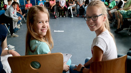 Schüler sitzen im Stuhlkreis in einem Schulzimmer. Zwei Schülerinnen haben sich in Richtung der Kamera umgedreht und lächeln.