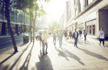 Im Sonnenschein laufen mehrere Personen über den Bürgersteig einer Geschäftsstraße, vorbei an den Schaufenstern der Geschäfte.