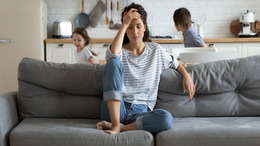 Eine Frau sitzt auf dem Sofa und fasst sich erschöpft, mit geschlossenen Augen, an die Stirn. Im Hintergrund toben ihre beiden Kinder durch die Küche des Hauses.