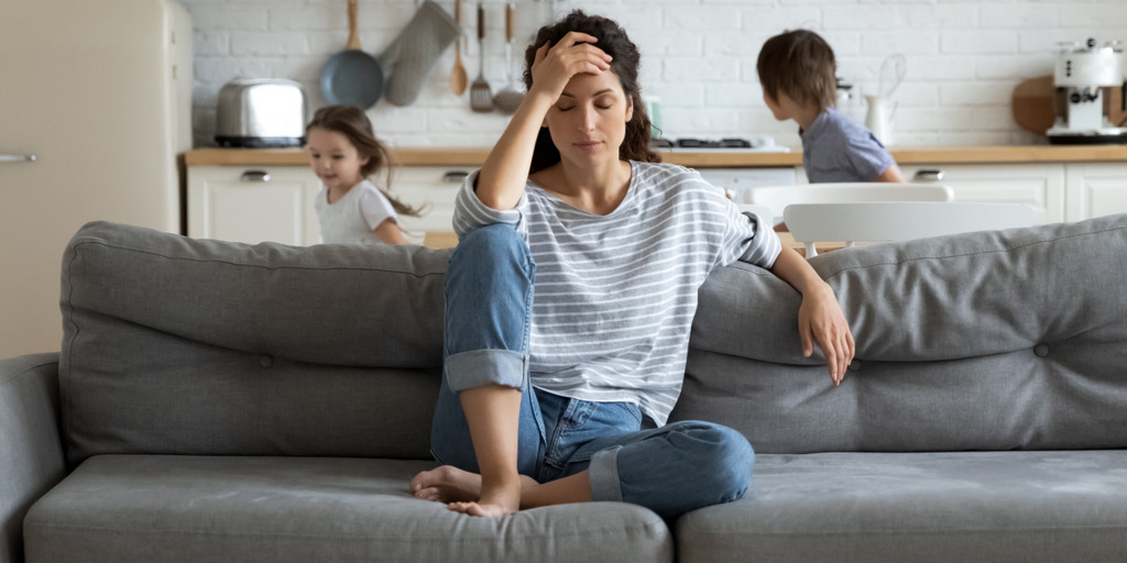 Eine Frau sitzt auf dem Sofa und fasst sich erschöpft, mit geschlossenen Augen, an die Stirn. Im Hintergrund toben ihre beiden Kinder durch die Küche des Hauses.