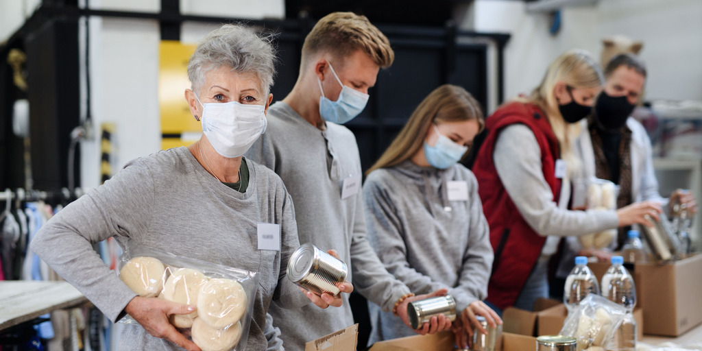 Mitarbeiterinnen und Mitarbeiter einer wohltätigen Organisation stehen nebeneinander hinter einem langen Tisch und packen Wasserflaschen, Konservendosen und eingeschweißte Brötchen in Kartons. Eine ältere Mitarbeiterin ist im Vordergrund zu sehen und blickt in die Kamera.