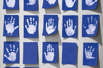 Das Bild zeigt blaue Klebezettel, die geordnet an einer Wand kleben. Auf den Zetteln sind mit weißer Farbe die Abdrücke von Kinderhänden zu sehen.