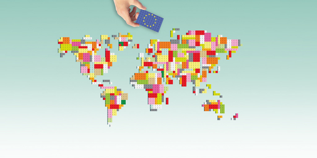 Weltkarte aus Legosteinen dargestellt. Oberhalb der Karte ist eine Hand mit einem Stein, der mit einer EU-Flagge bedruckt ist, dargestellt