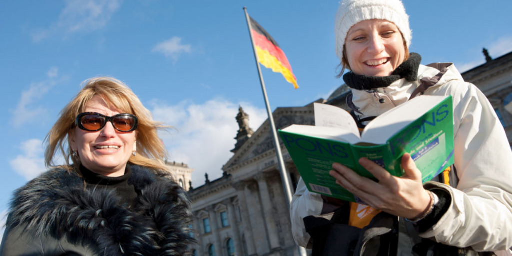 Zwei Frauen, die Deutsch als Fremdsprache lernen, stehen vor dem Reichstagsgebäude in Berlin und lächeln in die Kamera. Eine der Frauen hält ein aufgeschlagenes Deutsch-Lehrbuch in der Hand.