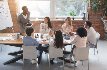 Teammeeting im Loftbüro: Acht Personen sitzen um einen Holztisch und applaudieren einem stehenden Kollegen mit Tablet; im Hintergrund Whiteboard mit Diagrammen, große Fenster und Zimmerpflanzen.