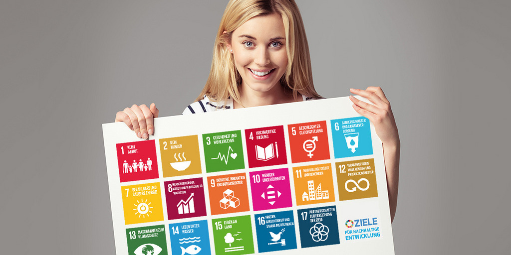Eine blonde, junge Frau hält ein Plakat. Auf dem Plakat werden die 17 Icons der Nachhaltigkeitsziele dargestellt.