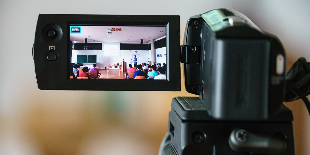 Über den Bildschirm einer schwarzen Videokamera sieht man ein Klassenzimmer. An der Leinwand steht ein Lehrer, vor ihm sitzen Schüler:innen