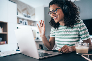 Junge Frau sitz lachend und winkend vor einem Laptop an einem Tisch. Sie trägt Kopfhörer und ist vermutlich in einem Onlinetermin