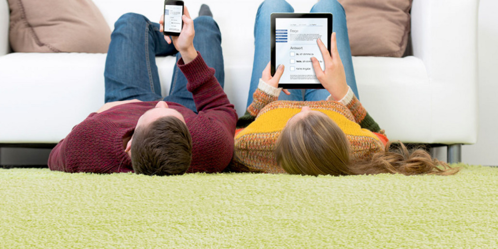 Ein junger Mann und eine junge Frau liegen nebeneinander auf dem Rücken vor ihrem Sofa, auf das sie ihre Beine abgelegt haben. Der Mann benutzt auf einem Smartphone den Wahl-O-Mat, die Frau macht dasselbe auf einem Tablet-Computer.
