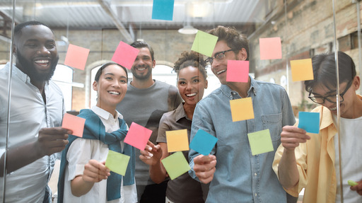 Eine Gruppe von jungen, lachenden Menschen klebt Post its an eine Glasscheibe