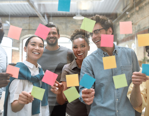 Eine Gruppe von jungen, lachenden Menschen klebt Post its an eine Glasscheibe