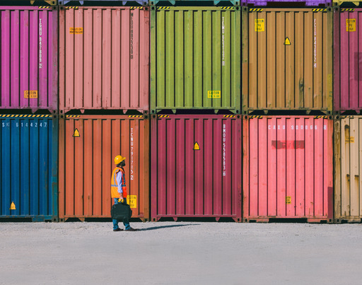 Several colorful shipping containers are stacked in neat rows, with a person in a safety vest and helmet walking across the wide, empty ground in front.