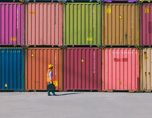 Several colorful shipping containers are stacked in neat rows, with a person in a safety vest and helmet walking across the wide, empty ground in front.
