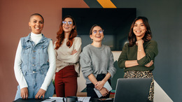 Erfolgreiche Unternehmerinnen, die fröhlich lächelnd hinter einem Tisch in einem Sitzungssaal stehen. Gruppe von multikulturellen Geschäftsfrauen, die in einem modernen Büro zusammenarbeiten
