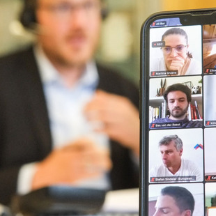 [Translate to English:] Der Trainer spricht vor dem Bildschirm, im Vordergrund ein Handy mit Gesichtern der Teilnehmenden