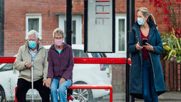 An elderly couple is sitting on the bench of a bus stop. Another lady is standing next to them a few meters away.