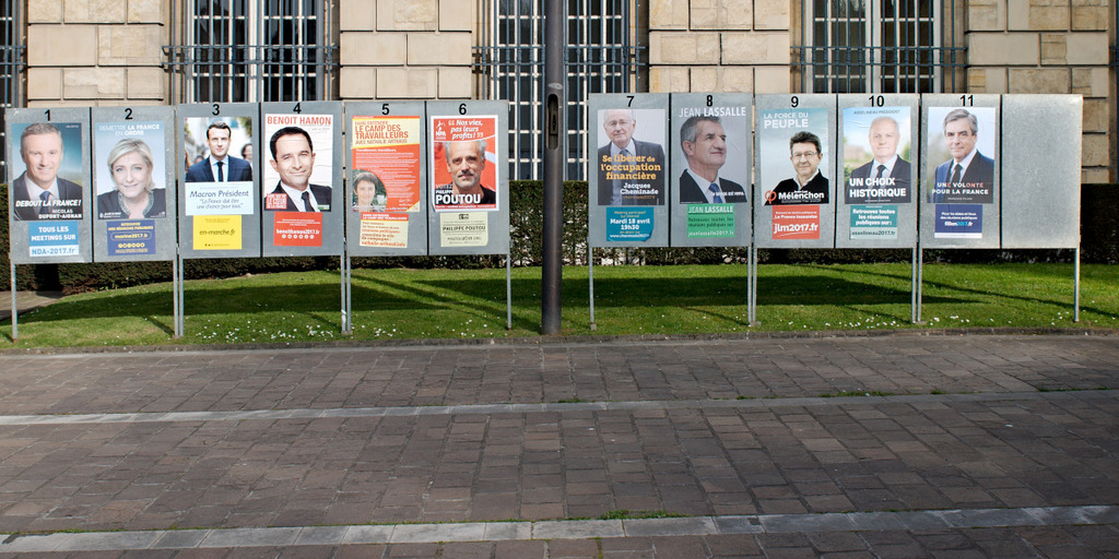 Plakatständer aus Metall, beklebt mit 11 Plakaten, stecken im Rasen vor einem Gebäude. Auf den Plakaten sind die Kandidaten für die französische Präsidentschaftswahl 2017 zu sehen.
