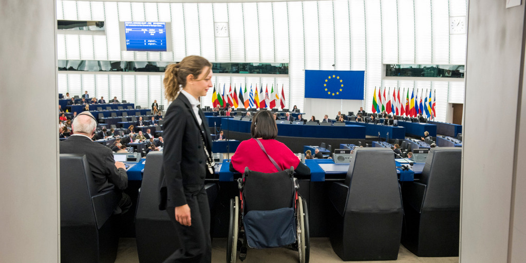 Blick von einem Gang in den Plenarsaal des EU-Parlaments in Straßbourg. Das Parlament ist voll besetzt; im Bildvordergrund sitzen zwei Parlamentarier und eine Parlamentsmitarbeiterin läuft durchs Bild.