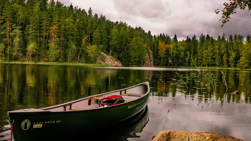 Das Bild zeigt ein ruhiges Gewässer, an dessen Ufer ein grünes Kanu vertäut ist. Im Hintergrund erstreckt sich ein dichter Wald mit überwiegend grünen Bäumen, von denen einige bereits herbstliche Verfärbungen aufweisen. Der Himmel ist bewölkt.