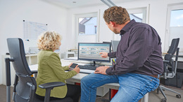 Eine Frau und ein Mann sitzen an und auf einem Schreibtisch in einem Büro, blicken auf einen PC-Bildschirm und diskutieren.