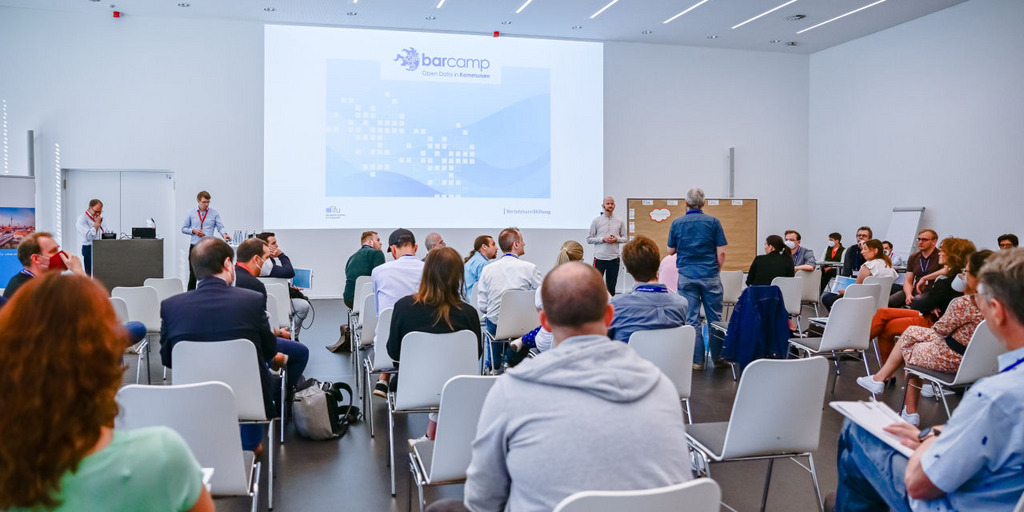 [Translate to English:] Blick von hinten im Raum auf die Teilnehmerinnen des Open-Data-BArcamps 2021 in Bochum. Vorne sind die Moderatoren und Gastgeber und eine Begrüßungsfolie ist an die Wand geworfen.