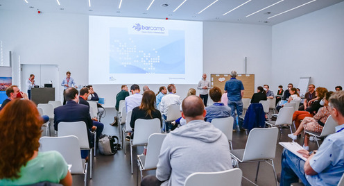 Blick von hinten im Raum auf die Teilnehmerinnen des Open-Data-BArcamps 2021 in Bochum. Vorne sind die Moderatoren und Gastgeber und eine Begrüßungsfolie ist an die Wand geworfen.