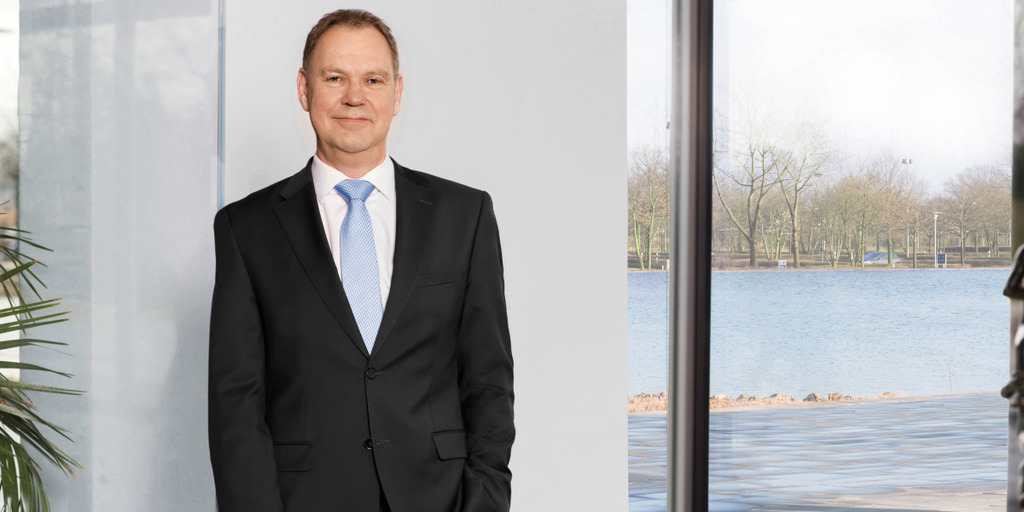 Aart De Geus, Vorstandsvorsitzender der Bertelsmann Stiftung, steht in einem Raum vor einer weißen Wand. Links von ihm eine Palme, rechts eine Glasfront, durch die der Stiftungsee zu sehen ist.