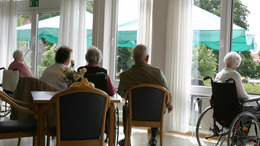 In einem Pflegeheim sitzen einige Seniorinnen und Senioren in einem Aufenthaltsraum und blicken hinaus in den sonnenbeschienenen Park.
