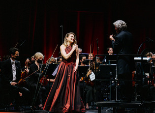 Francesca Pia Vitale, Siegerin der NEUEN STIMMEN 2022, steht während der NEUEN STIMMEN 2022 auf der Bühne der Stadthalle in Gütersloh in einem roten Kleid und singt. Neben ihr steht der Dirigent Jonathan Darlington, der die hinter ihr spielenden Duisburger Philharmoniker dirigiert.