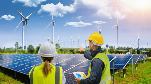 Eine Frau und ein Mann stehen vor eine Solaranlage. Im Hintergrund sieht man mehrere Windräder.