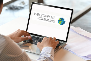 Auf einem Laptop sieht man das Logo der Wissensplattform Weltoffen Kommune. Vor dem Laptop sitzt eine Person.