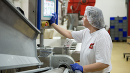 Eine Mitarbeiterin einer Lebensmittelfabrik trägt Arbeitskleidung, Handschuhe und ein Haarnetz und steht an einer Maschine, die sie über einen daran befestigten Touchscreen bedient.