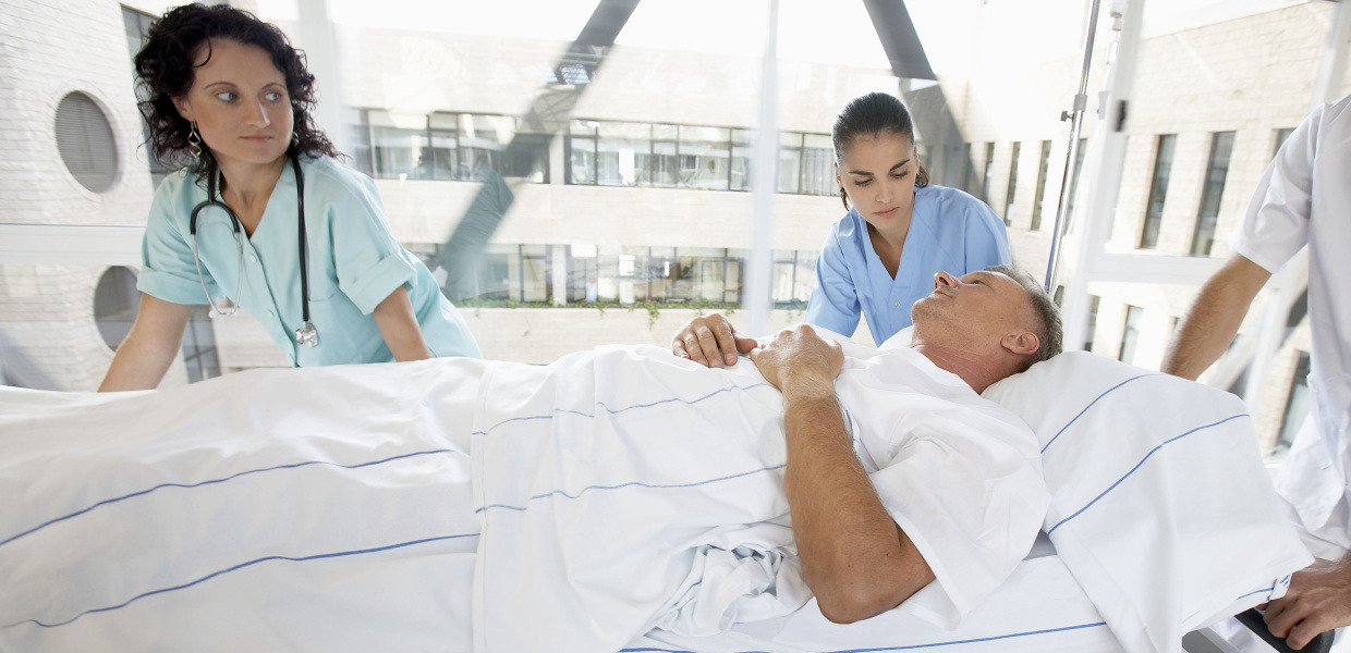 Ein Mann wird in einem Krankenbett von zwei Mitarbeiterinnen eines Krankenhauses geschoben.