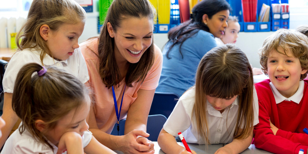 Eine Lehrerin sitzt mit mehreren Schülern an einem Tisch und bearbeitet Aufgaben. Im Hintergrund eine weitere Lehrerin mit Schülern.