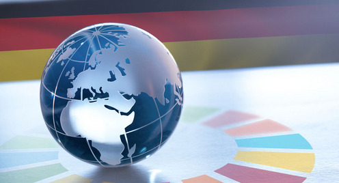 Eine Weltkugel aus Glas liegt auf einem Untergrund, auf dem strahlenförmig von ihm das Logo für die Nachhaltigkeitsziele der Vereinten Nationen ausgeht. Im Hintergrund sieht man die Farben der deutschen Flagge.