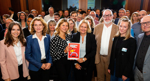 Group picture of German Federal Minister of the Interior Nancy Faeser, Bertelsmann Stiftung Executive Board member Daniela Schwarzer and members of the Citizens' Assembly during the event at which the "Forum against Fakes" were submitted. Nancy Faeser and a member of the Citizens' Assembly are surrounded by the other participants and present the recommendations to the camera.