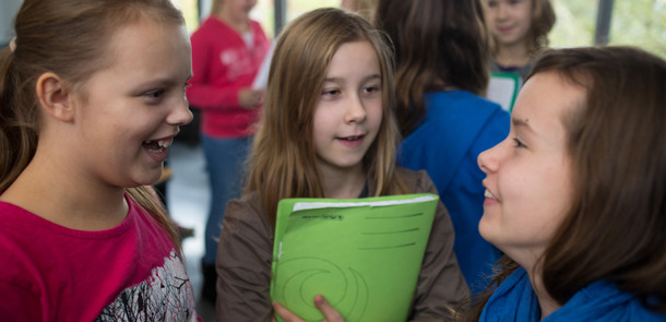 Drei Mädchen stehen zusammen und reden miteinander, eine hält eine grüne Mappe an ihre Brust gedrückt